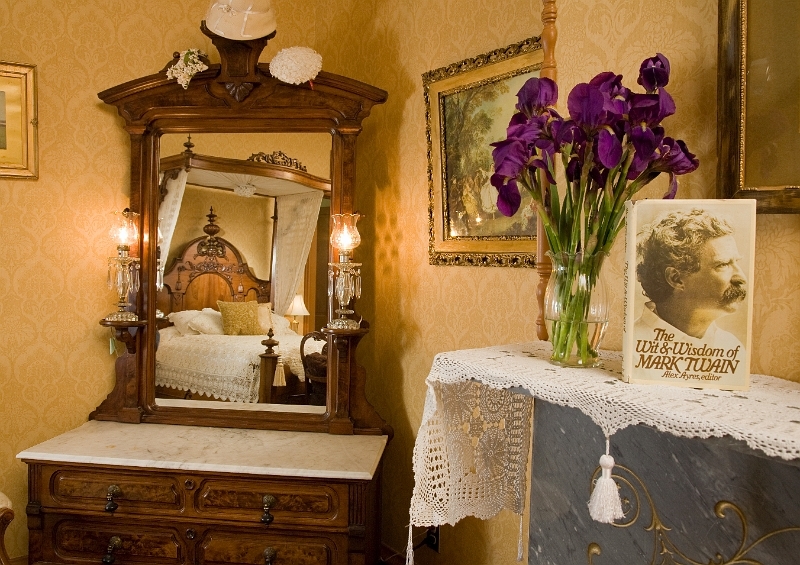 A vintage dresser with a mirror reflecting a four-poster bed, a vase of purple flowers, and framed artwork on the wall.