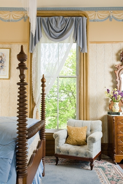 A corner of a bedroom with a four poster bed, a window with sheer curtains, and an armchair.