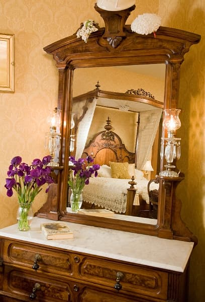 A vintage dresser with a mirror reflecting a four-poster bed, a vase of purple flowers, and framed artwork on the wall.