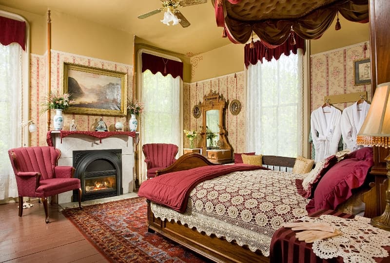 A luxurious bedroom with a four-poster bed, a cozy fireplace, plush armchairs, and a window with sheer curtains.