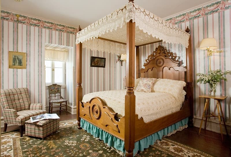 A bedroom with a large four-poster bed, a sitting area with an armchair and ottoman, and striped wallpaper.