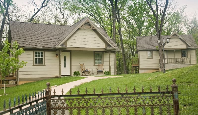 Two charming cottages with inviting front porches, set on a tree-lined property with a decorative fence.