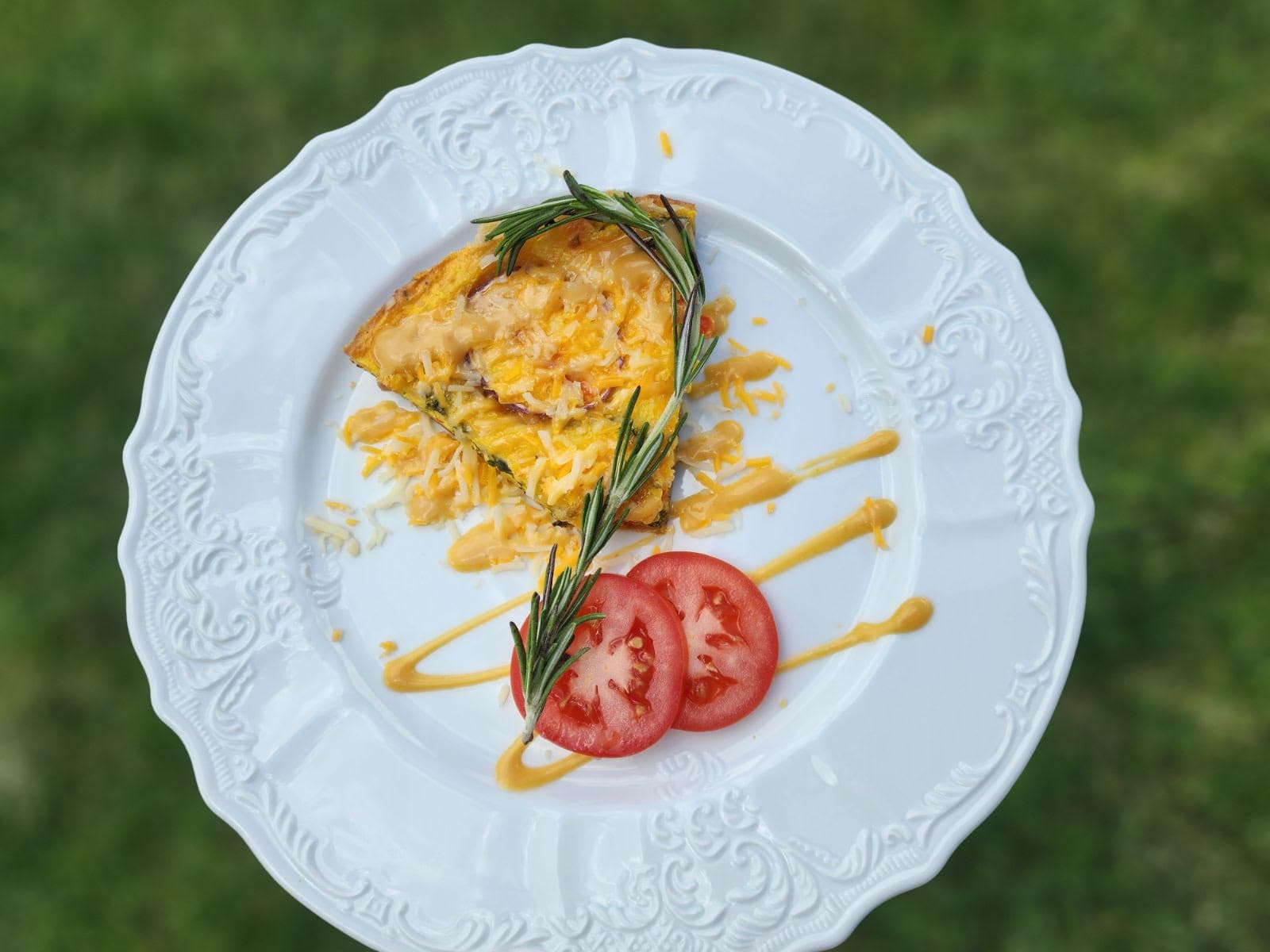 A slice of cheesy frittata garnished with fresh herbs and accompanied by sliced tomatoes on a decorative plate.