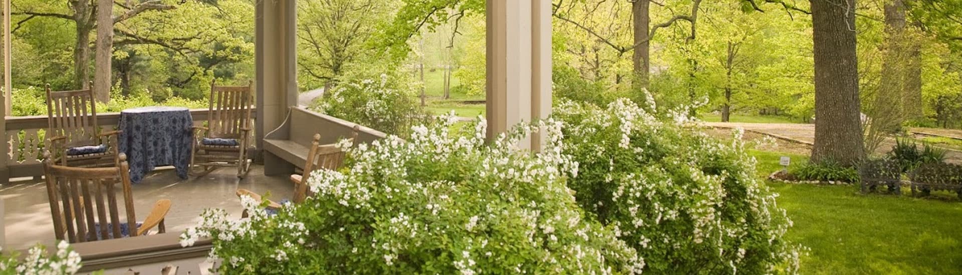 A peaceful porch surrounded by lush greenery and blooming white flowers offers a tranquil outdoor seating area.
