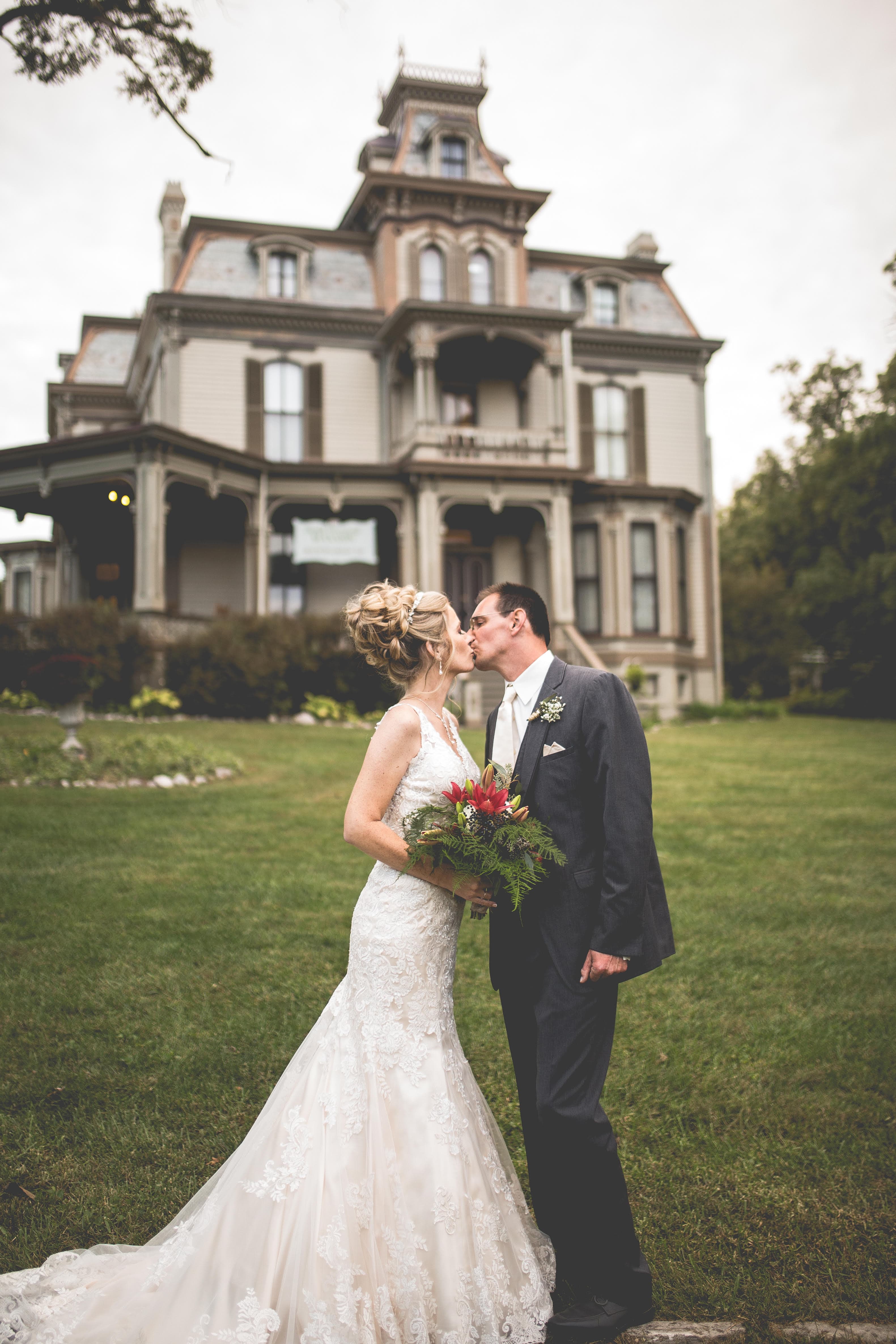 A bride and groom kiss in front of a beautiful Victorian house while holding a bouquet.