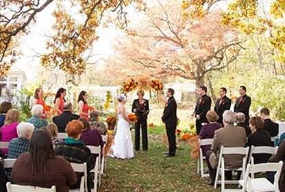 An outdoor wedding ceremony under a canopy of autumn trees, with guests seated and the couple exchanging vows.