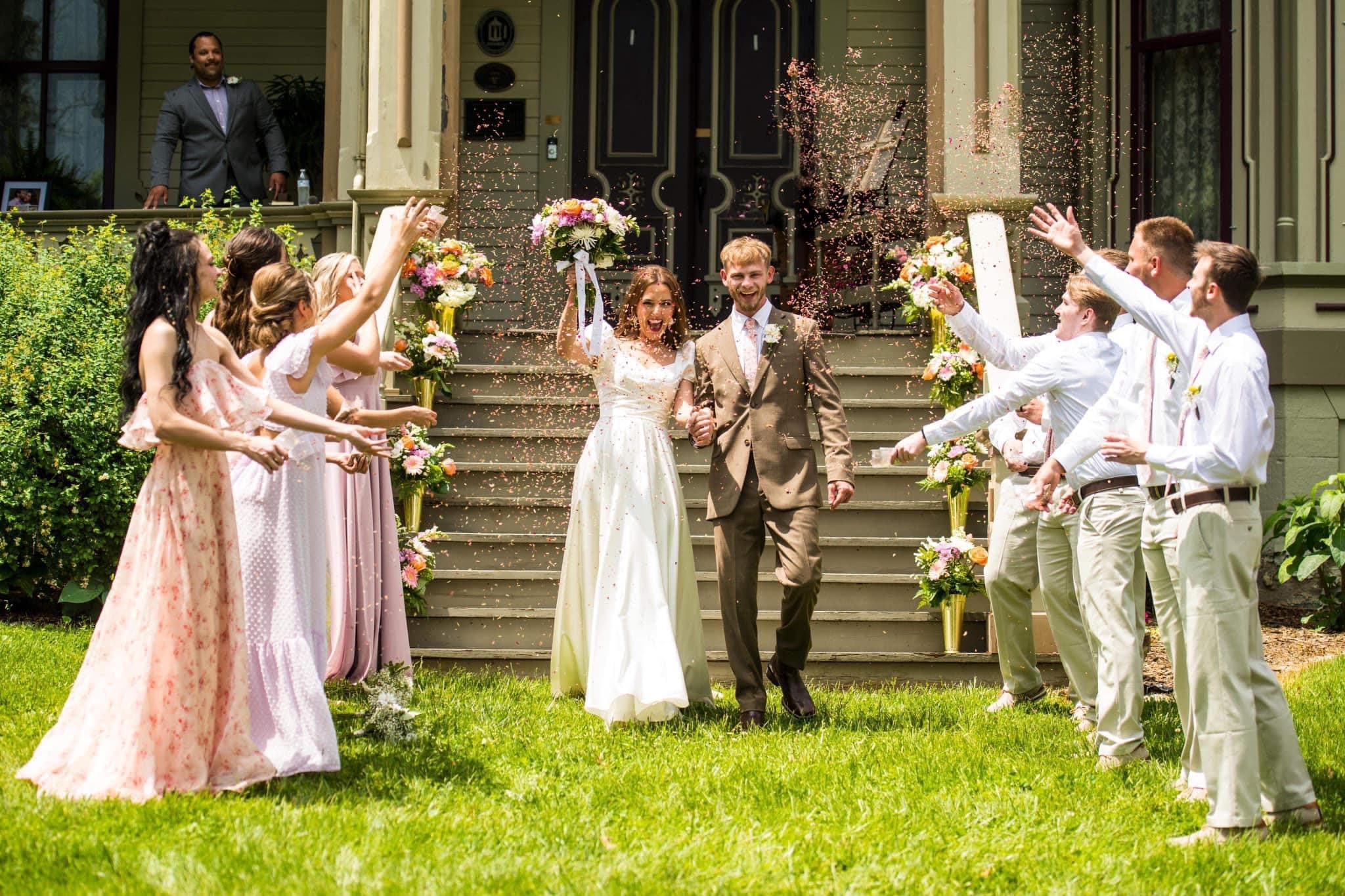 A joyful newlywed couple exits a house while guests shower them with confetti and flowers.