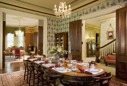 An elegant dining room with chandeliers, vintage decor, and a beautifully set long table. The adjacent room enhances the space.