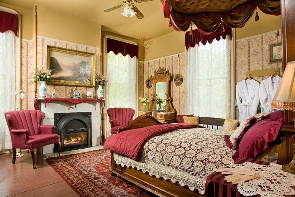 A Victorian-style bedroom featuring a cozy fireplace, elegant decor, and a luxurious bed with lace bedding and rich red accents.